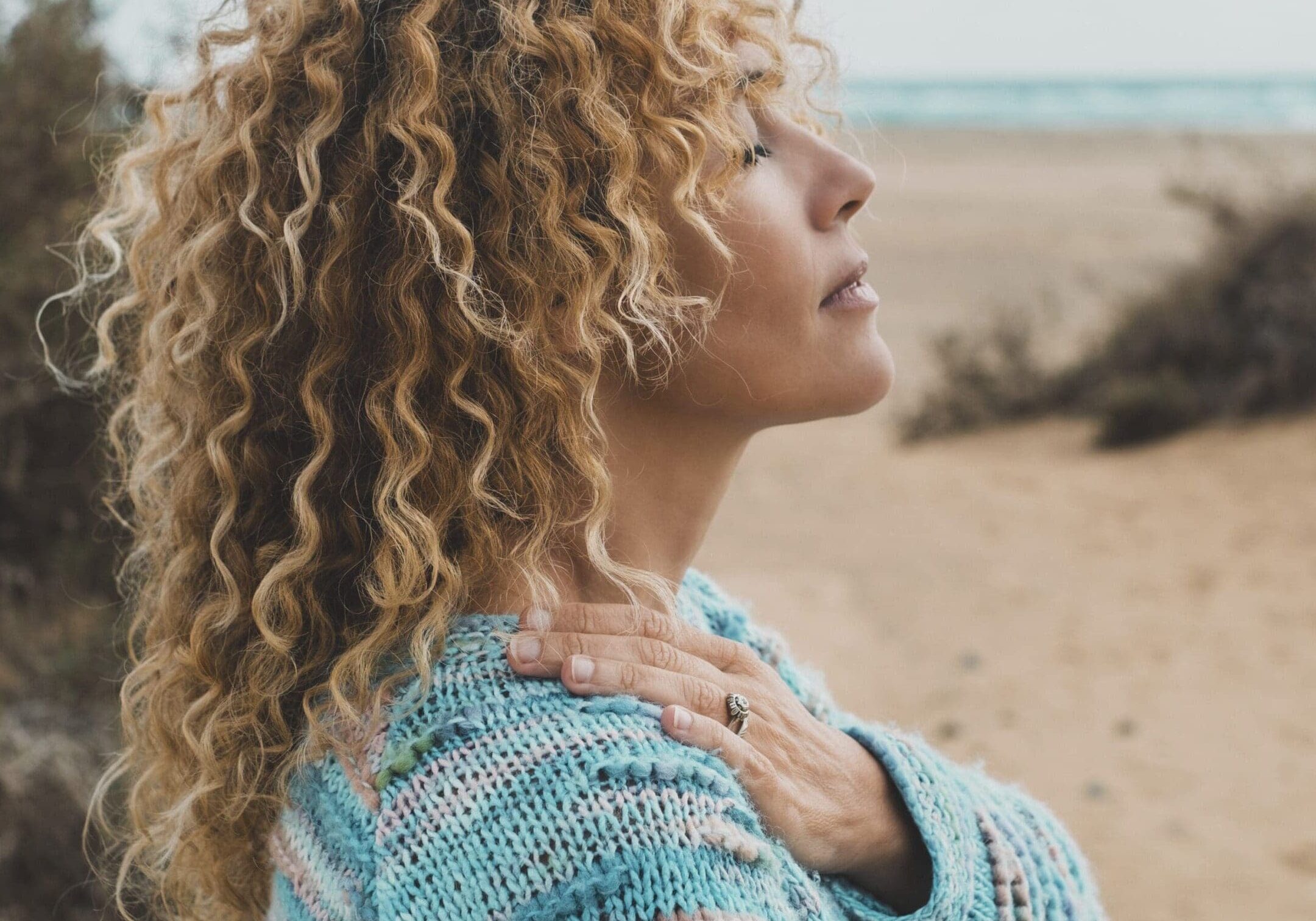 Woman with curly hair in a sweater, standing on a beach with eyes closed.
